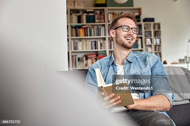 laughing man sitting with book on couch in the living room - young man relaxing on sofa stock pictures, royalty-free photos & images