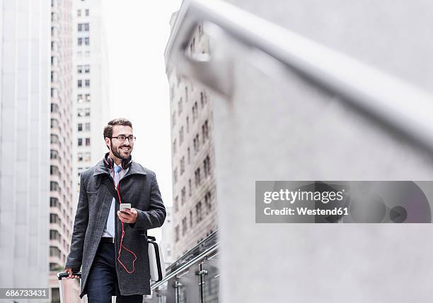 usa, new york city, smiling businessman with cell phone and headphones on the go - trolley stock-fotos und bilder