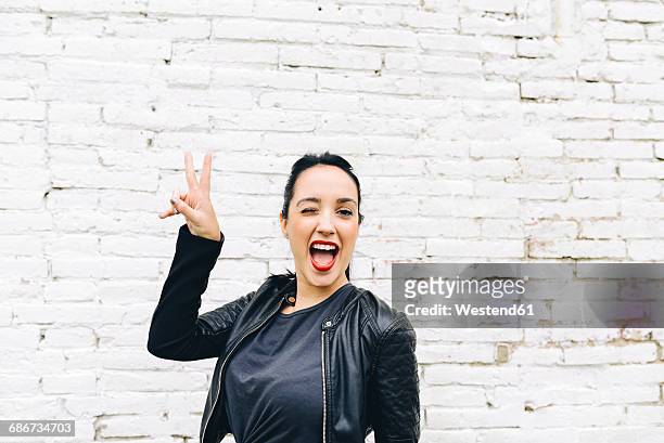 portrait of young woman in front of brick wall making victory sign - peace sign gesture stock-fotos und bilder