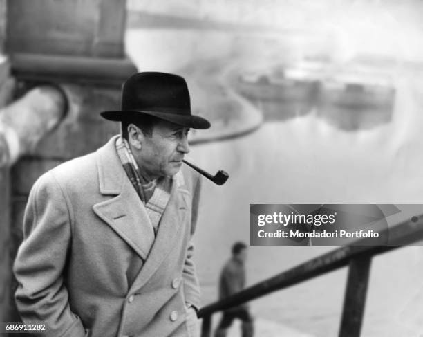 Belgian writer Georges Simenon, smoking a pipe, climbing the metal stairs on the docks of Porta Ticinese. In the background, in the mist, there are...