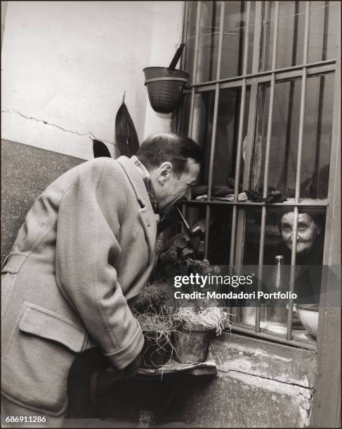 Belgian writer Georges Simenon, smoking a pipe, bowing to look through a barred window, beyond which we see the face of an elderly woman. Milan, 1957