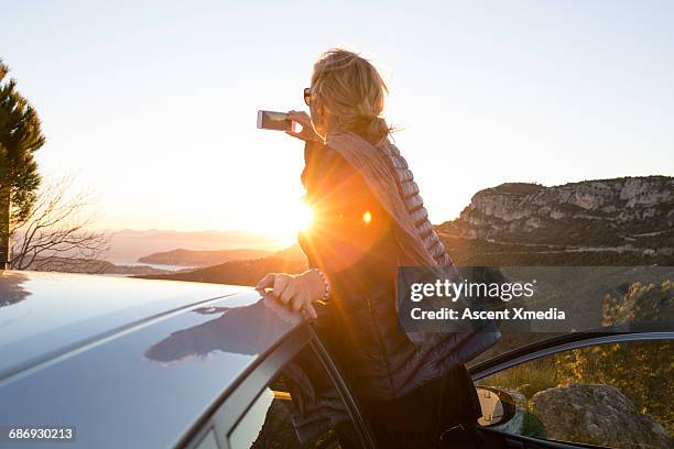 woman takes picture of sunrise over sea, hills - autofoto stockfoto's en -beelden