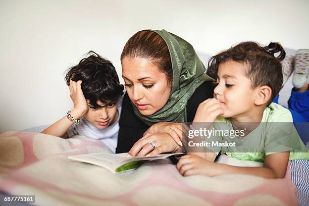 mother reading book to boys while lying on bed at home - headscarf home stockfoto's en -beelden