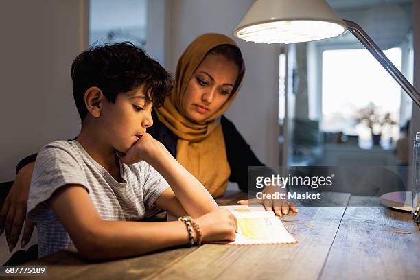 Mother and son reading book under illuminated desk lamp at home