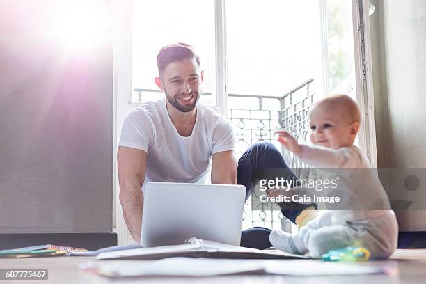 father working on laptop and watching baby daughter playing on floor - leanintogether stock pictures, royalty-free photos & images