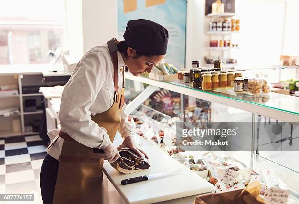 female owner cutting meat at counter in grocery store - loja de conveniencia imagens e fotografias de stock