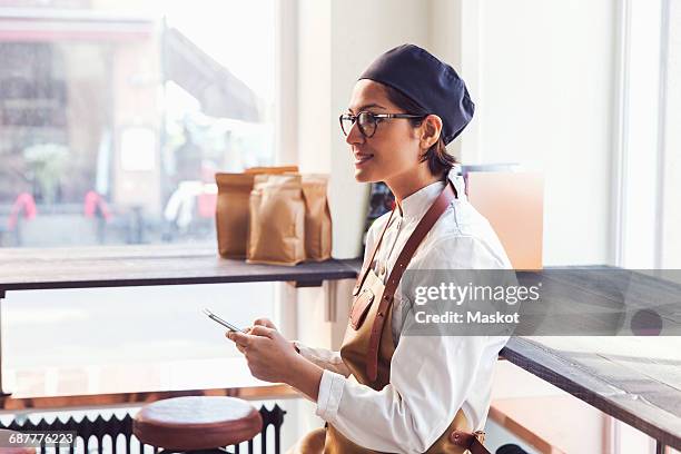 smiling female owner holding smart phone while standing by window in grocery store - mercante foto e immagini stock