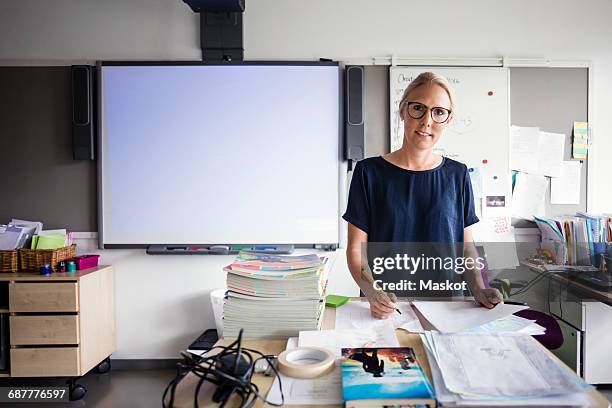 portrait of teacher with papers against blank whiteboard in classroom - blank book on desk stock-fotos und bilder