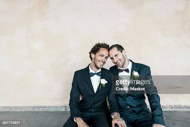 Portrait of newlywed gay couple sitting on bench against wall