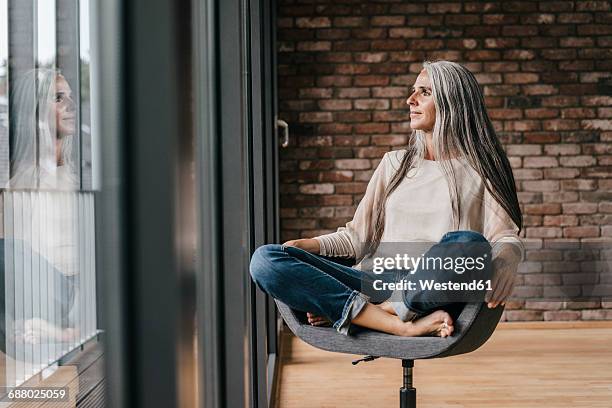 woman with long grey hair sitting on chair at the window - schneidersitz stock-fotos und bilder