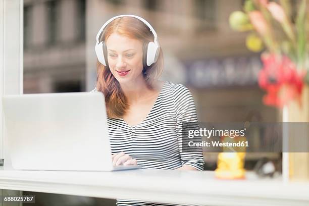 smiling woman sitting in a coffee shop using headphones and laptop - achterdeur stockfoto's en -beelden