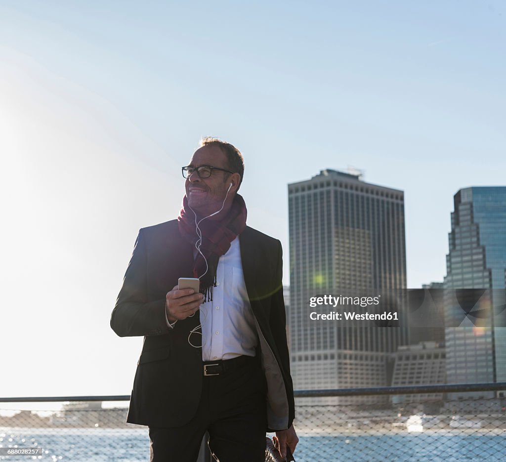 USA, Brooklyn, smiling businessman with earphones and smartphone in front of Manhattan skyline