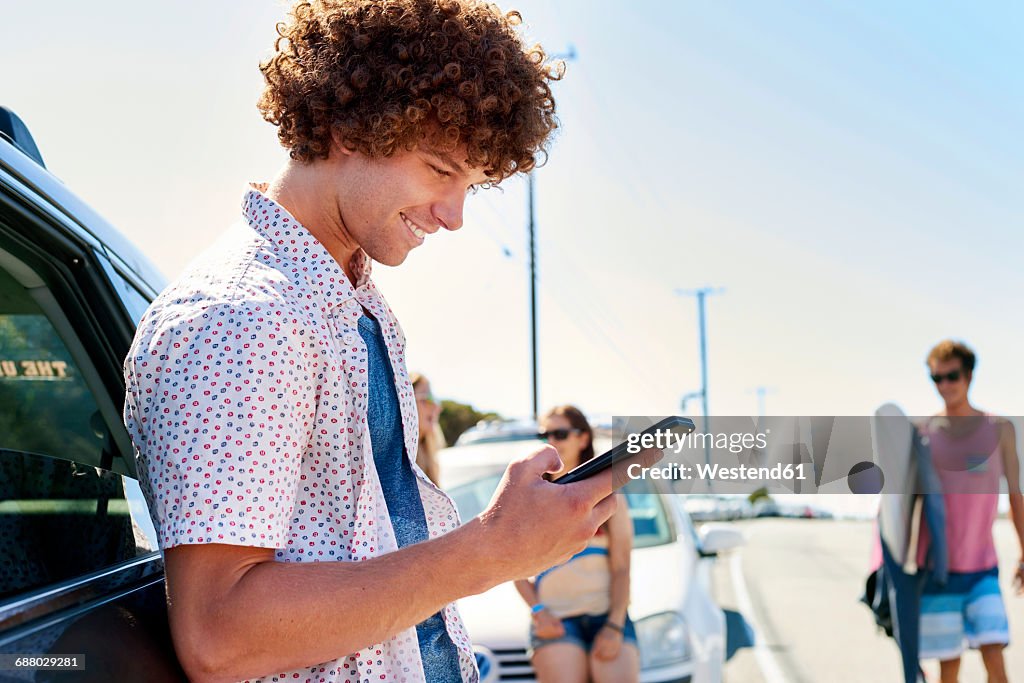 Smiling young man at a car checking his cell phone