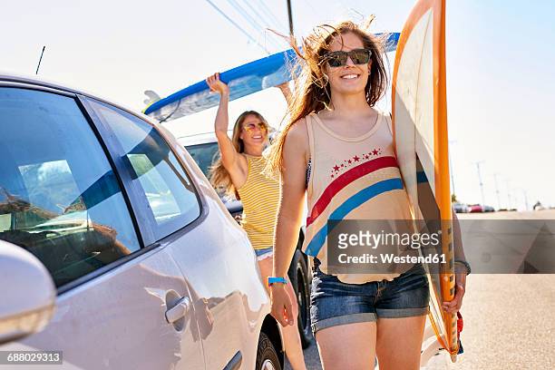 two smiling young women carrying surfboards on coastal road - californie surf stock pictures, royalty-free photos & images