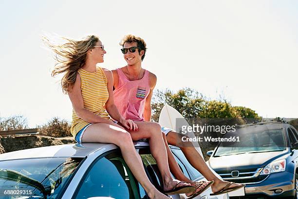 smiling young couple sitting on car roof - sitting on surfboard ストックフォトと画像