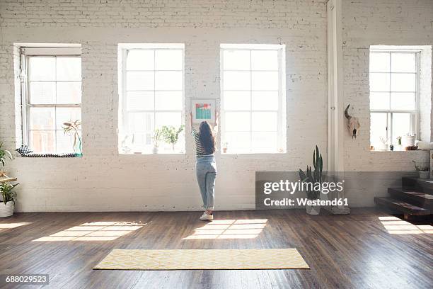 young woman attaching picture frame to brick wall in a loft - loft brique photos et images de collection