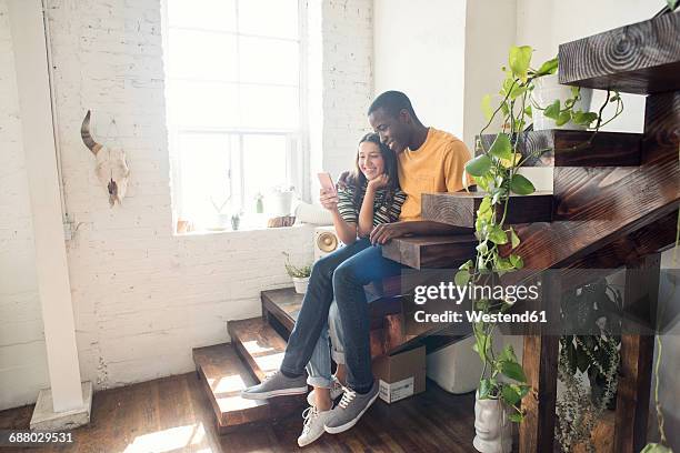smiling young couple sitting on stairs in a loft sharing cell phone - apartamento tipo loft fotografías e imágenes de stock