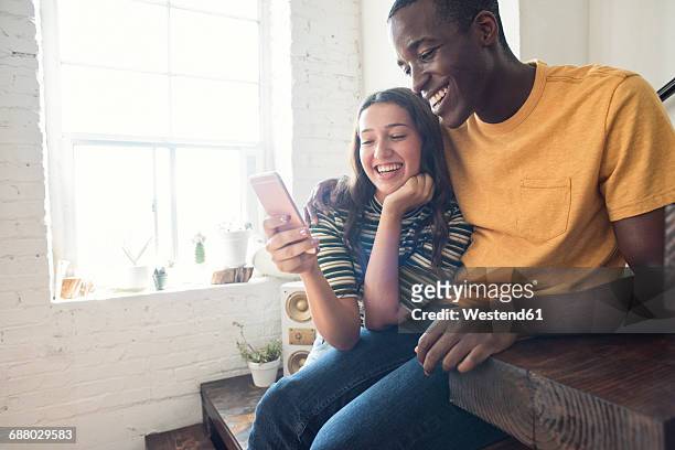 happy young couple sitting on stairs in a loft sharing cell phone - happy couple using cellphone stockfoto's en -beelden