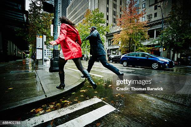 couple jumping over puddle on city street - レインコート ストックフォトと画像