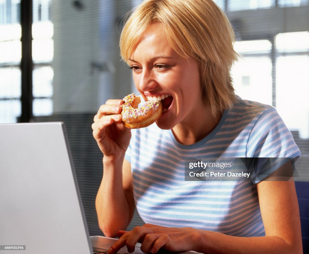 Young woman sitting eating doughnut at desk