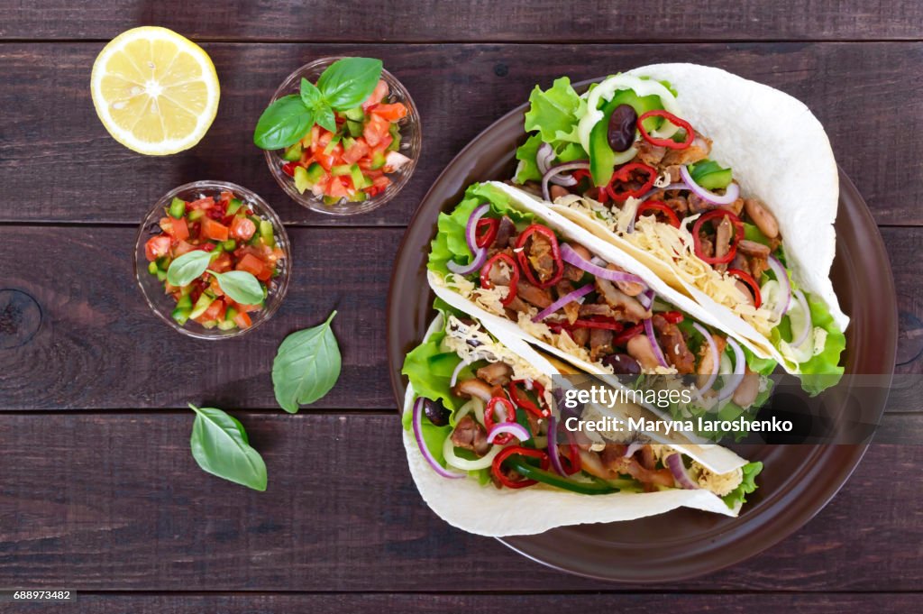 Tacos is a traditional Mexican dish. Tortilla stuffed with chicken, bell and hot peppers, beans, lettuce, cheese, blue onion with salsa sauce on dark wooden background. Top view.