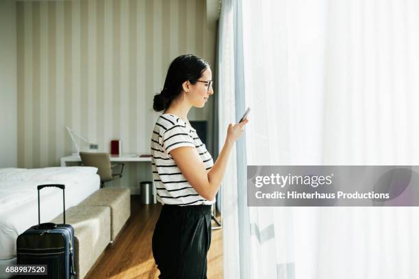 young woman using her phone by hotel room window - curtain hotel stock-fotos und bilder