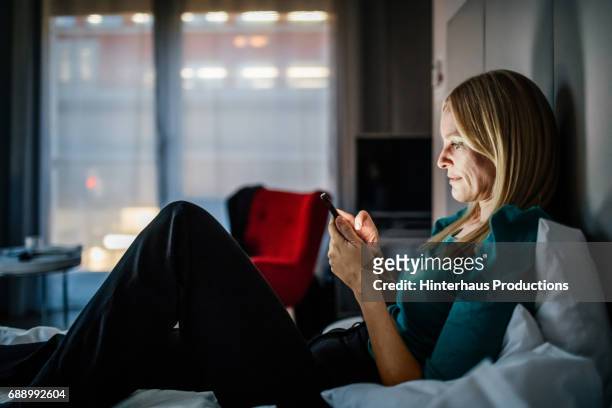 businesswoman reclining on bed using smartphone - slaapkamer zijaanzicht stockfoto's en -beelden