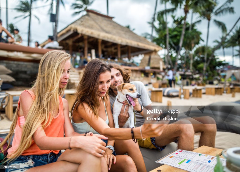 Group of friends taking selfies - two girls, one guy  and a cute dog with them at the beach cafe