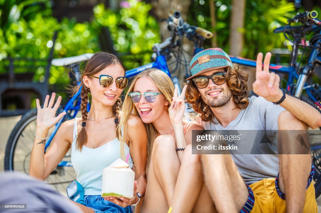 Grupo de amigos - dos chicas y un hombre - sentado en la arena, con descanso en la sombra de los árboles. Pequeño perro cerca. Bicicletas detrás de ellos