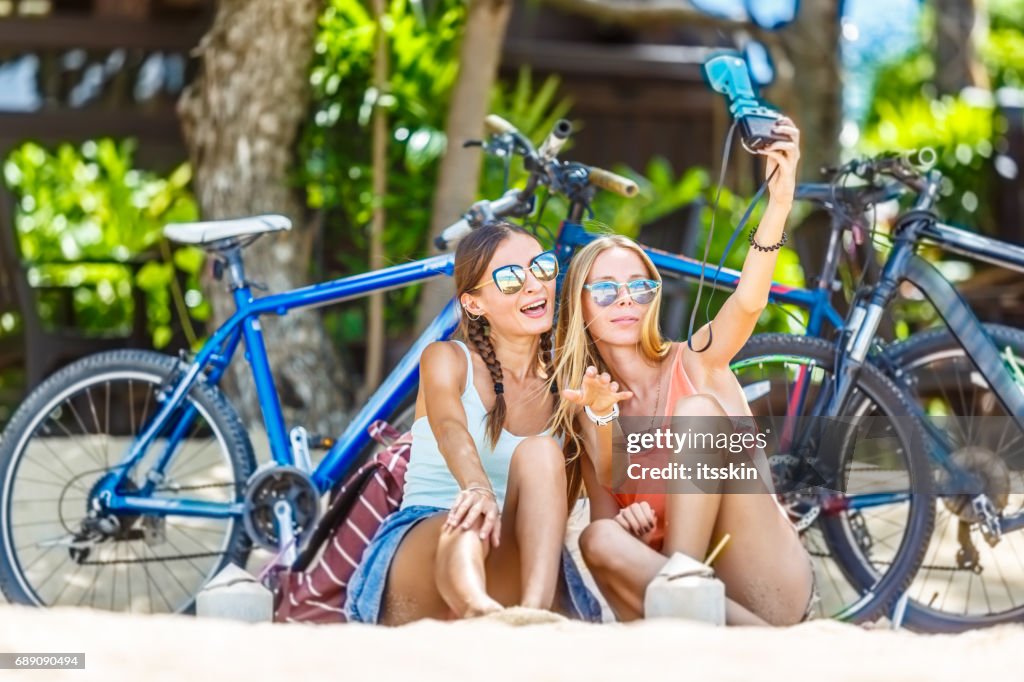 Two girlfriends sitting on sand, having rest in the shade of trees, taking selfies. Bicycles behind them