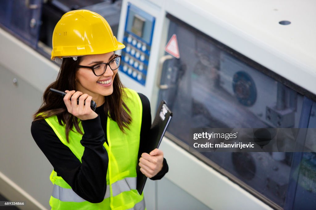 Jonge vrouw in fabriek met gele helm en wifi-telefoon