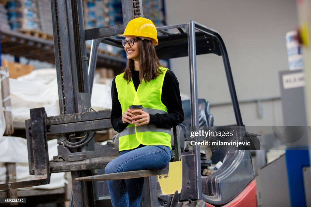 Beautiful girl at factory in a vest and helmet sitting on forklift.