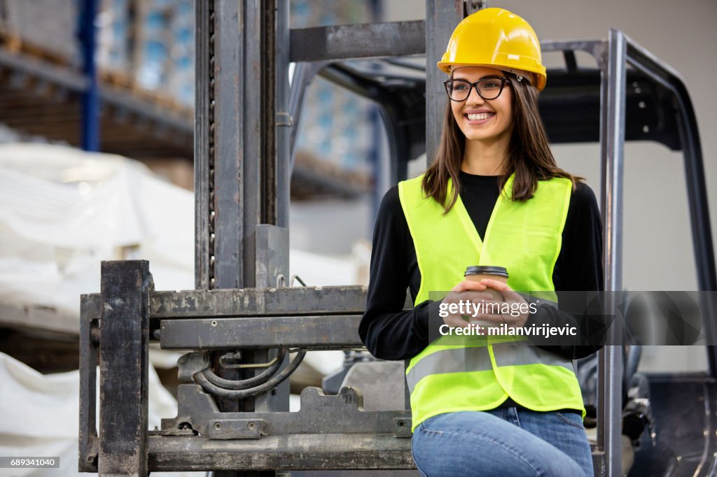 Beautiful girl at factory in a vest and helmet near forklift with coffee.