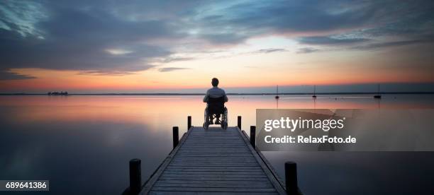 senior mann sitzt im rollstuhl am ende des see steg beobachten majestätische wolkengebilde in der abenddämmerung - romantischer sonnenuntergang stock-fotos und bilder