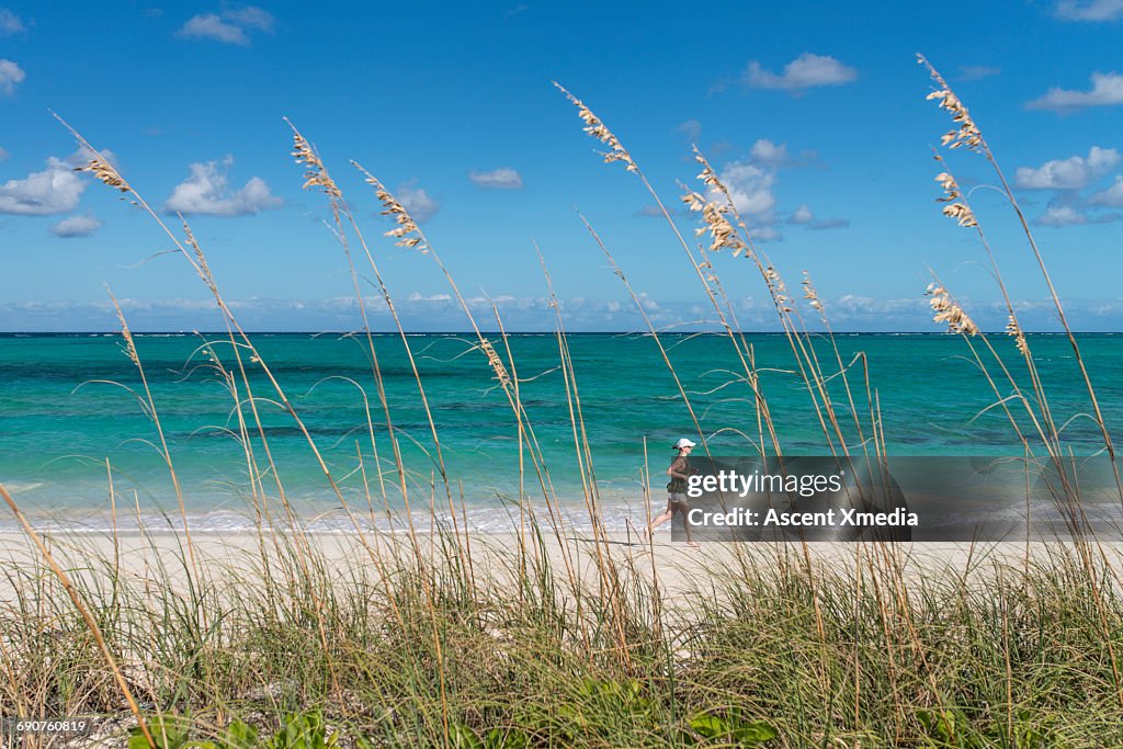 Distant woman runs along sand beach, past grasses