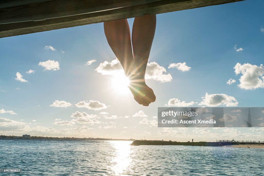 Woman's feet dangle above calm water, seaside