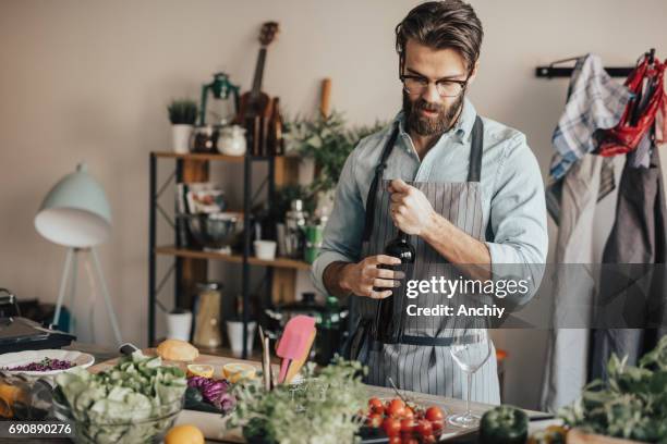 handsome man in kitchen opening a bottle of red wine - hipster in a kitchen stock pictures, royalty-free photos & images