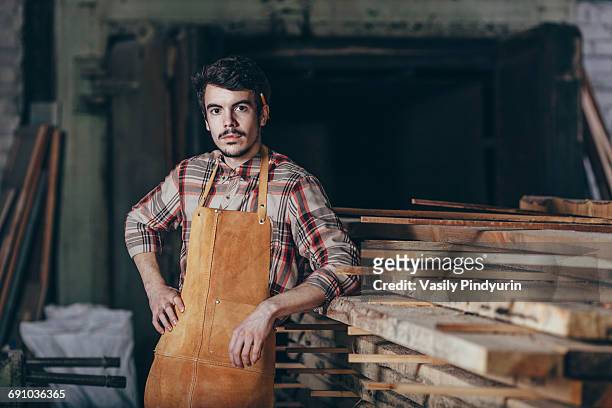 portrait of carpenter standing by timber stack in workshop - carpenter stockfoto's en -beelden