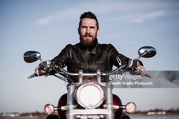 portrait of confident biker sitting on motorcycle against sky - motociclista fotografías e imágenes de stock
