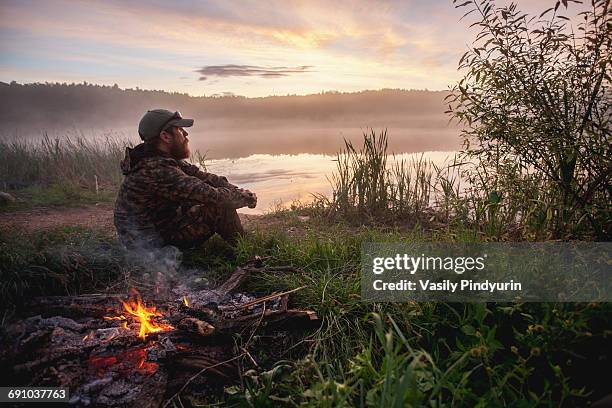 side view of hunter sitting by bonfire on field at lakeshore during sunset - hunting stock pictures, royalty-free photos & images