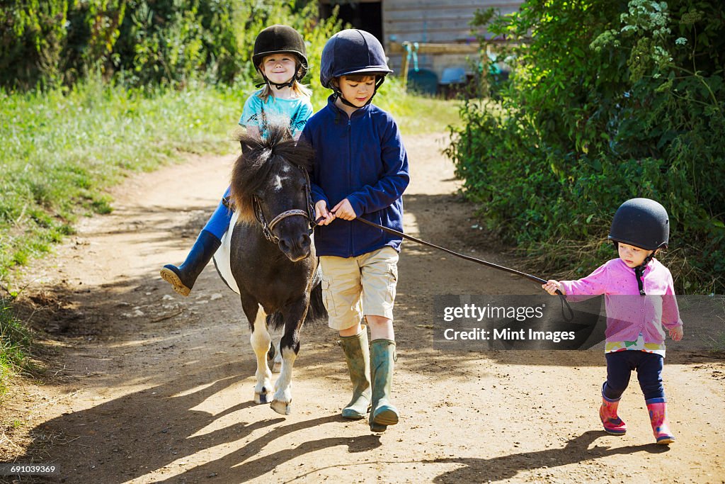 A boy, a toddler, and a girl riding a pony on a dirt path.