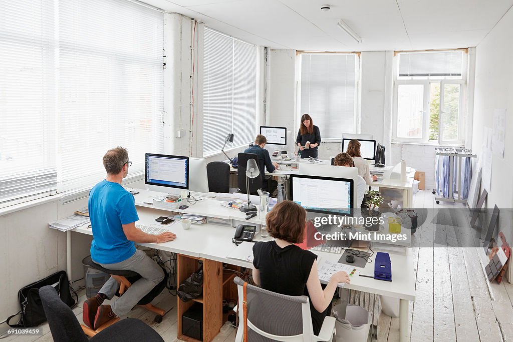 A modern office, workstations for staff. Elevated view of six people seated at desks. A man using an ergonomic kneeler chair.