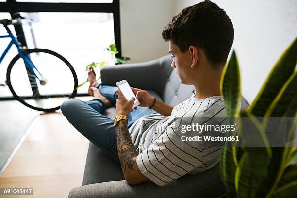 teenage boy sitting on couch at home using cell phone - teen boy barefoot stock-fotos und bilder