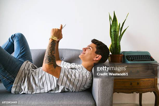 smiling teenage boy lying on couch at home taking selfie with cell phone - typewriter stockfoto's en -beelden