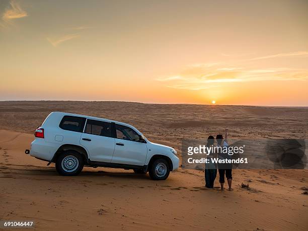 oman, al raka, two young women standing besides off-road vehicle on a desert dune watching sunset - oman landscape stock pictures, royalty-free photos & images