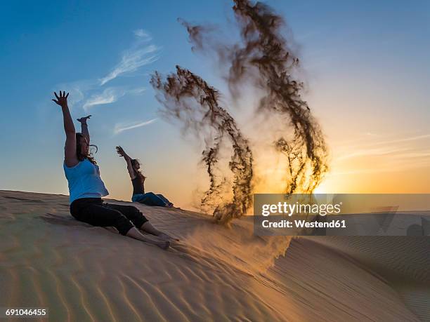 oman, al raka, two young women sitting on a dune in rimal al wahiba desert throwing sand in the air - middle east friends stock pictures, royalty-free photos & images