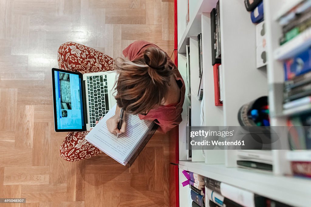 Woman at home sitting on floor using laptop and taking notes