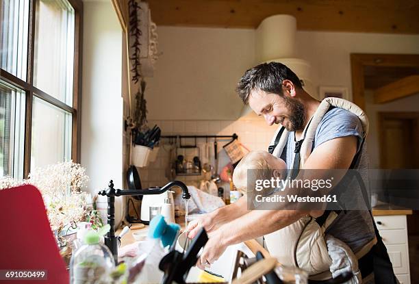 happy father with baby in baby carrier doing the dishes - house husband stock pictures, royalty-free photos & images
