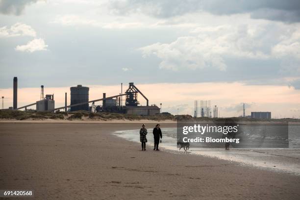 People walk dogs on Redcar Beach, in front of a backdrop of the shuttered Teesside Steelworks, formally operated by Sahaviriya Steel Industries Pcl,...