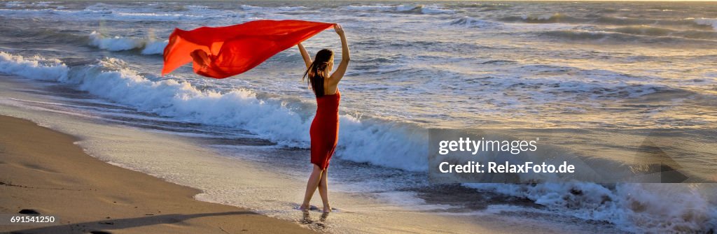 Mujer madura de pie en la playa del Mediterráneo sosteniendo la bufanda roja en el viento